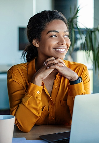 A woman in a yellow satin shirt is sitting at her laptop, looking out with her hands clasped under her chin. She is smiling and reassured