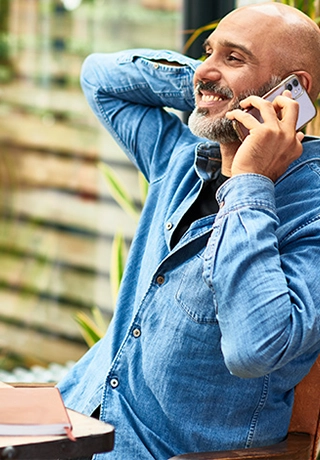 A man is sitting in a home conservatory type environment. He is wearing a blue shirt. He is on the phone. He is in a relaxed position, leaning back with his arm supporting his head