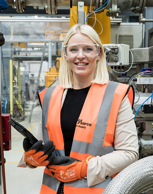 A blonde woman with a hi-vis jacket and safety glasses stands in a factory with a clipboard