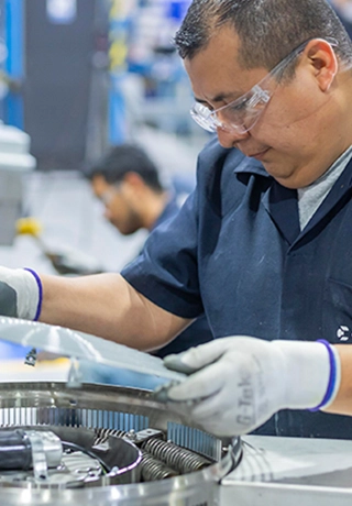 A senior manufacturing colleague stands over a stainless steel type cannister. He is wearing gloves and glasses and a short-sleeved navy blue shirt. 