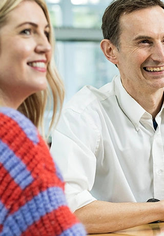A man and a woman are sitting together at a table smiling. The man is wearing a white Watson-Marlow shirt and the woman is wearing a red and blue striped jumper