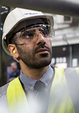 A man with a hi-vis jacket, hardhat and glasses stands in front of a pipe assessing it