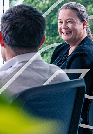 Two colleagues are talking to one another in front of a computer, one male, one female. The male has his back turned to the camera. The woman is looking at the screen with the man and smiling in agreement.