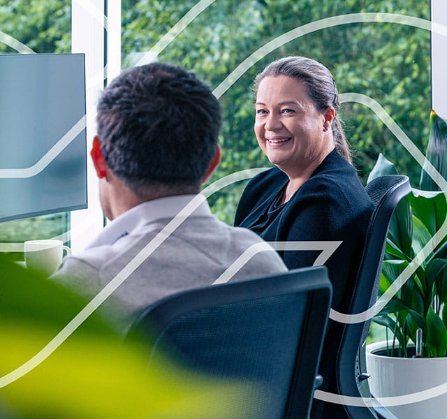 Two colleagues are talking to one another in front of a computer, one male, one female. The male has his back turned to the camera. The woman is looking at the screen with the man and smiling in agreement.