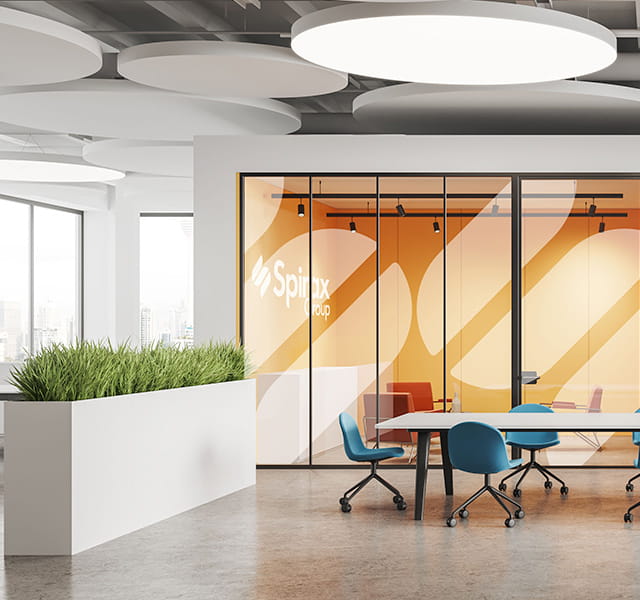 An office space with a table and blue chairs to the right and a tall-planter with green plants to the left. Behind the table and planter is an office-type room with the Spirax Group logo printed on the wall and two S icons covering the glass wall next to it