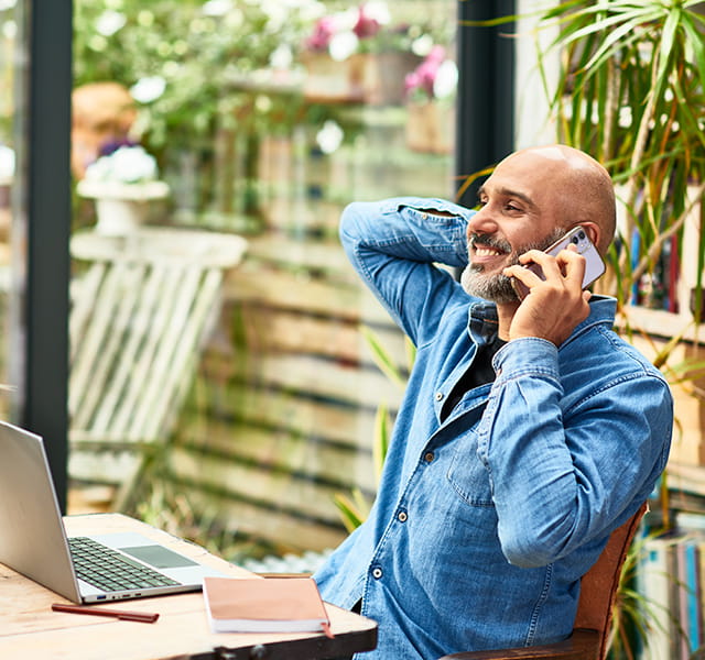 A man is sitting in a home conservatory type environment. He is wearing a blue shirt. He is on the phone. He is in a relaxed position, leaning back with his arm supporting his head