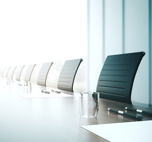 A long row of executive chairs at a board table. There are glasses of water at the table and a piece of white paper at each seat. The image fades from the right to the left