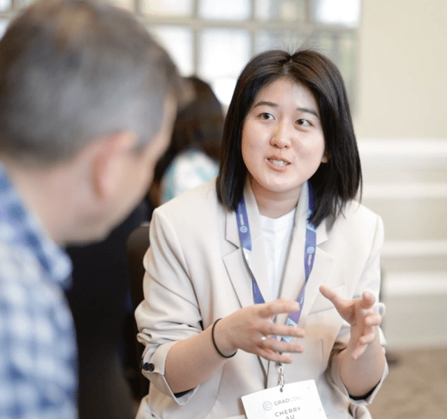 A woman wearing a cream blazer and a Company lanyard is talking to a male colleague wearing a blue checked shirt who is closer to the camera, out of focus and his face turned away