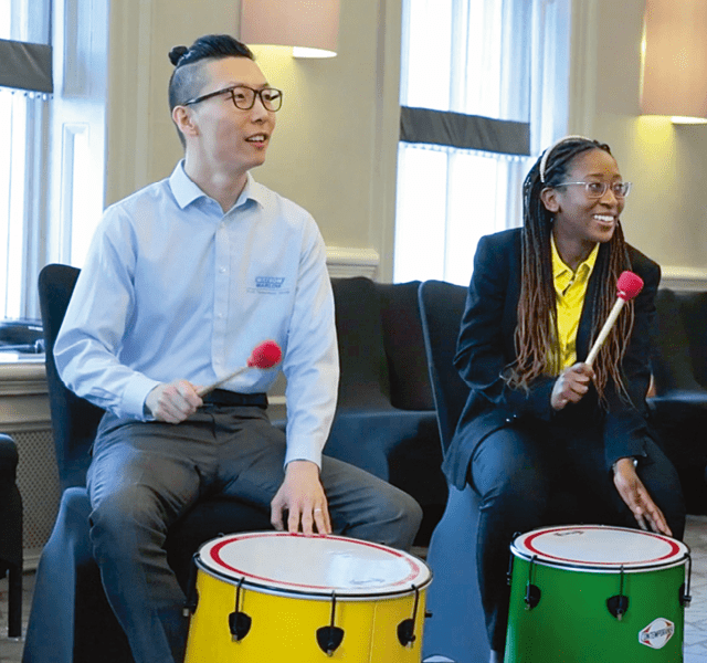 Two global graduates sitting down on chairs, playing a drum each at a wellbeing event