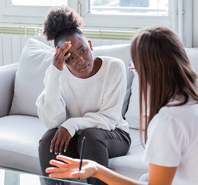 Two women are talking to one another on a sofa, deep in conversation. One is holding her head in one hand. The other has her face out of shot, but is holding her hand out to the side 