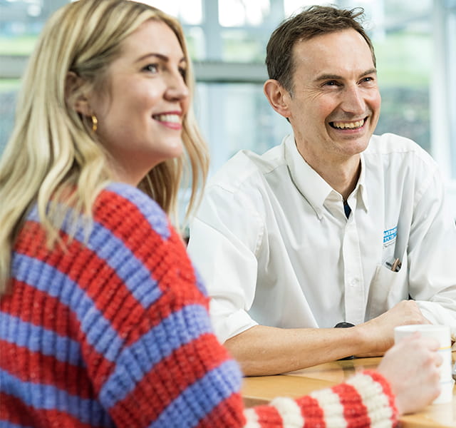 A man and a woman are sitting together at a table smiling. The man is wearing a white Watson-Marlow shirt and the woman is wearing a red and blue striped jumper