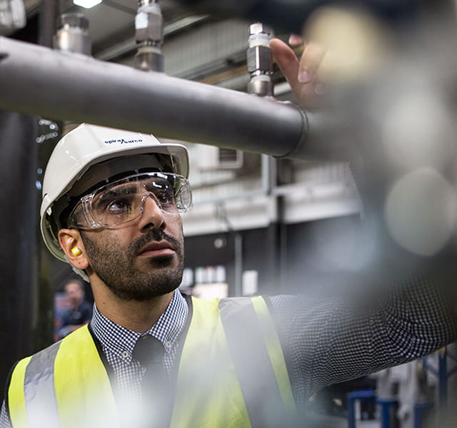 A man with a hi-vis jacket, hardhat and glasses stands in front of a pipe assessing it