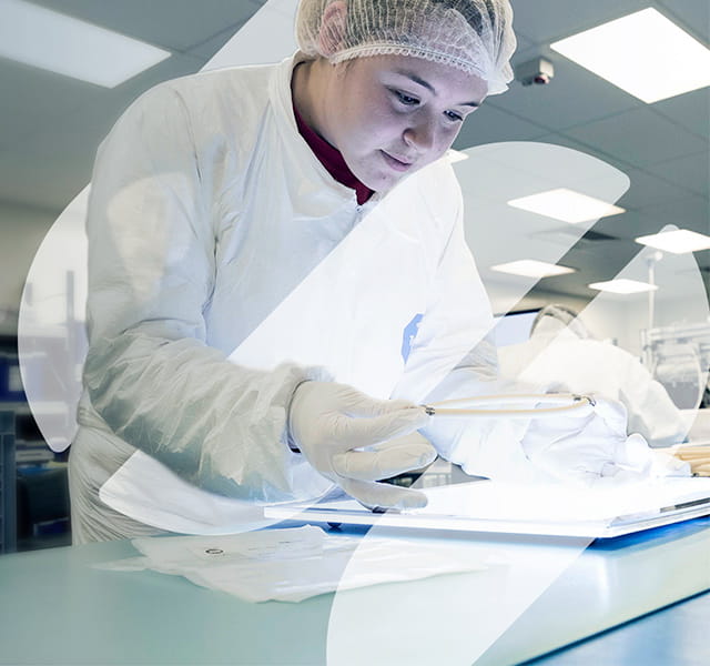 A woman leans over her experiment in a lab environment. She is wearing a hair net and a white pair of overalls and white gloves. There is the S shape for Spirax Group overlaid to the bottom right quarter of the image