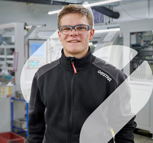 A male apprentice in a Gestra zip up hoodie stands in the manufacturing environment. He is smiling directly at the camera. 