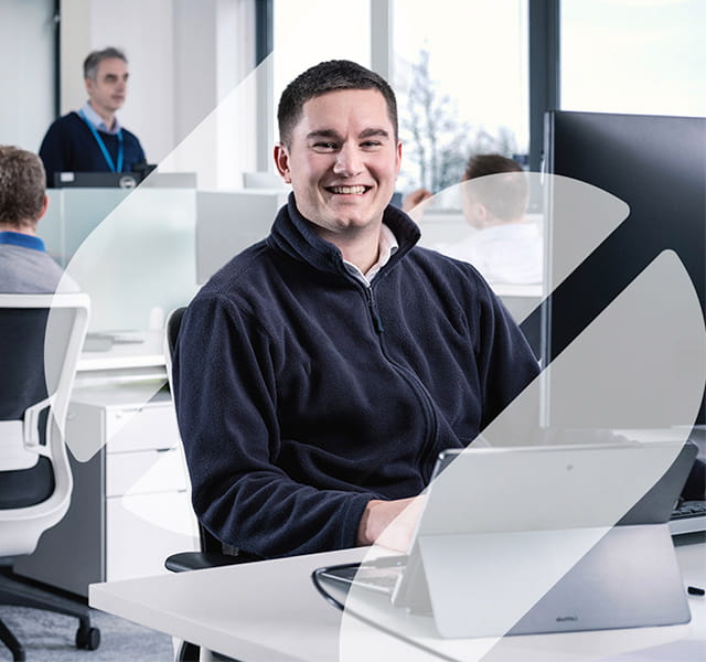 A close up of a man in an office sitting at his computer. He is looking at the camera directly and smiling. He is set within the Spirax Group S shape with him in the middle of it