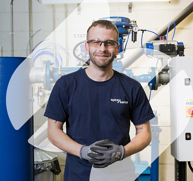 A man stands behind an engineered solution product. He is smiling directly at the camera wearing a Spirax Sarco t-shirt. He is wearing gloves and glasses. He is standing in the middle of the Spirax Group S shape overlaid on the image