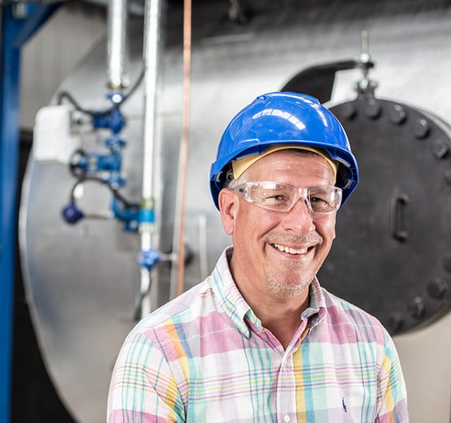 A Watson-Marlow colleague is pictured within a manufacturing environment. He is looking into the camera directly and smiling. He  is wearing glasses, a blue hard hat and a pastel checked shirt