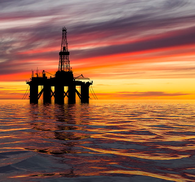 An oil platform in the middle of the sea at sunset. The sunset is pink and orange with dark grey clouds. 