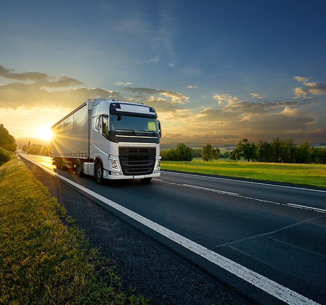 A lorry is driving down an empty road surrounded by fields. It is driving away from a sunset