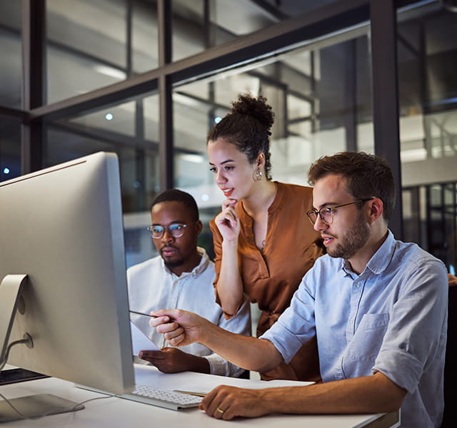 Two men and one women are huddled around a computer. One man is pointing at the screen. The woman is looking at the screen while holding her chin