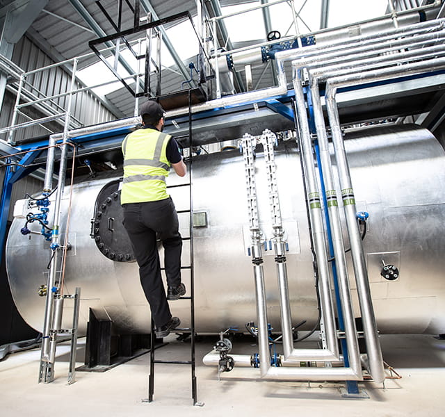 A man climbs a ladder wearing a hi-vis vest to access a higher platform on a boiler