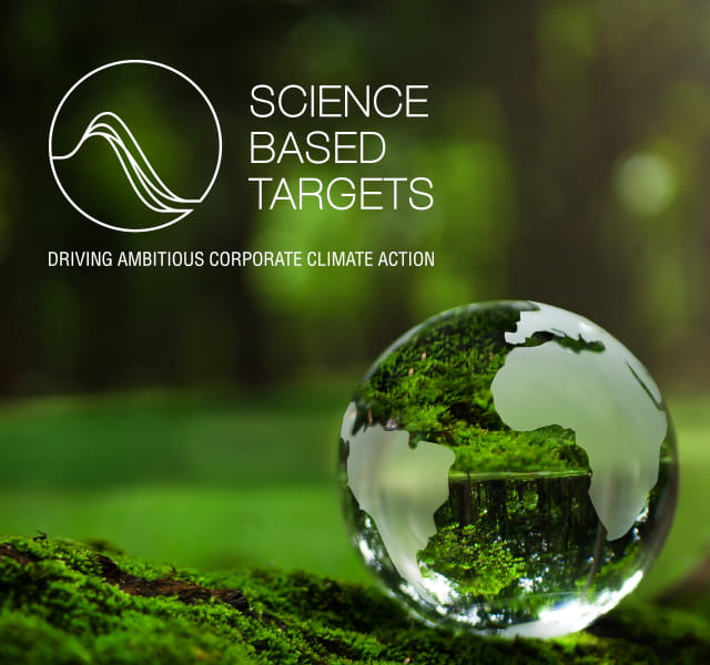 The SBTi logo is white and transparent. It is overlaid in the top left corner of the image. To the bottom right is a stylised clear globe which looks like it's made from a drop of water. The globe and the logo sit amongst an out of focus shot within a woodland environment