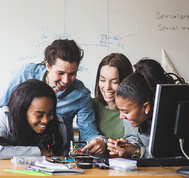 Four people are crowded around a fun electronic project set on a table in a learning environment