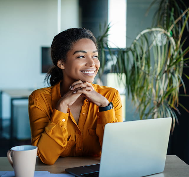 A woman in a yellow satin shirt is sitting at her laptop, looking out with her hands clasped under her chin. She is smiling and reassured