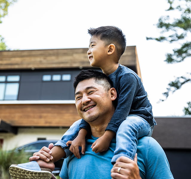 A man is walking away from a house with a little boy on his shoulders. They are both smiling