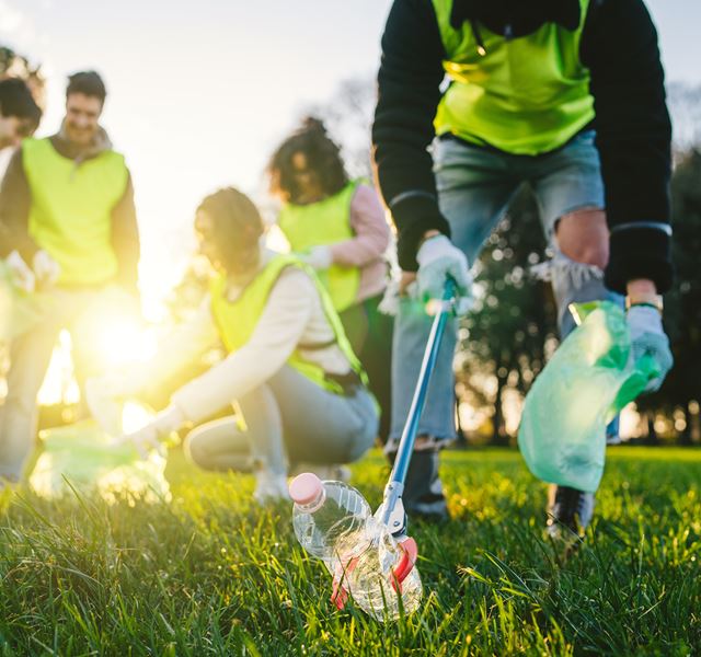 A group of people litter picking. They are wearing hi-vis jackets. Four people are in the background in discussion, a fifth person is closest to the camera and picking up a piece of rubbish with a grabber