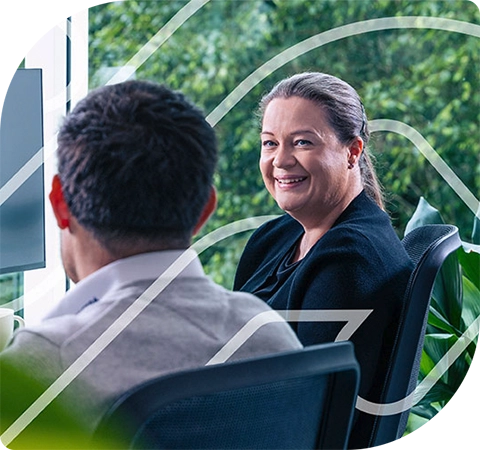 Two colleagues are talking to one another in front of a computer, one male, one female. The male has his back turned to the camera. The woman is looking at the screen with the man and smiling in agreement.