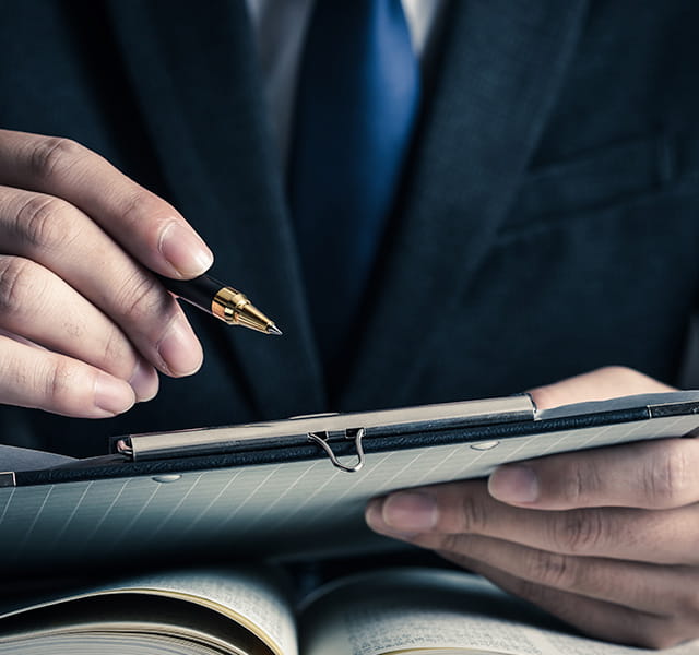 A close up shot of a person wearing a suit and blue tie. They have paper in their one hand and holding a pen with the other. 